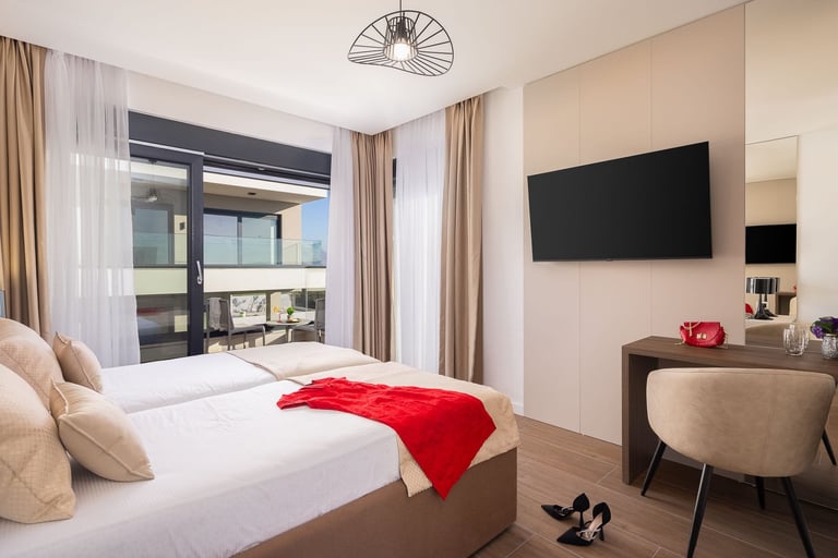 Modern hotel bedroom with white bedding, red throw blanket, wall-mounted TV, floor-to-ceiling windows with curtains, and contemporary furnishings