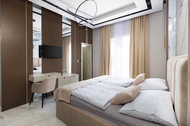 Modern bedroom with beige bed, white pillows, brown accent wall, built-in desk, floor-to-ceiling windows with beige curtains, and contemporary pendant lighting
