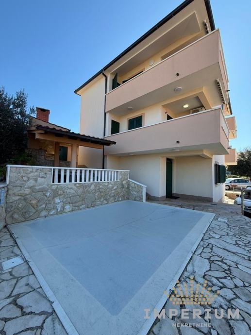 Modern three-story white residential building with stone driveway, terrace, and parked car under clear blue sky