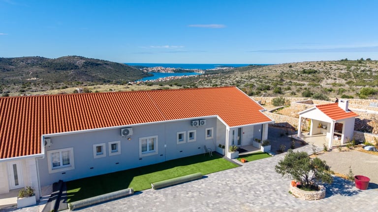 Pink residential house with orange roof and sea view, surrounded by Mediterranean landscape and sparse vegetation