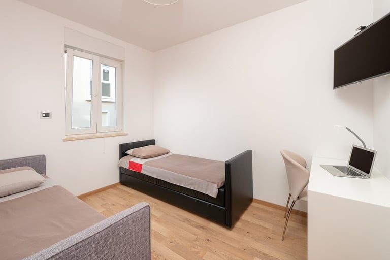 Modern dorm room with two beds, wooden floor, desk with laptop, window, and wall-mounted storage cabinet