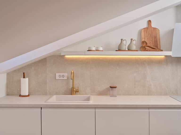 Modern minimalist kitchen with white countertop, stainless sink, gold faucet, and lit floating shelf displaying kitchen items and wooden cutting board