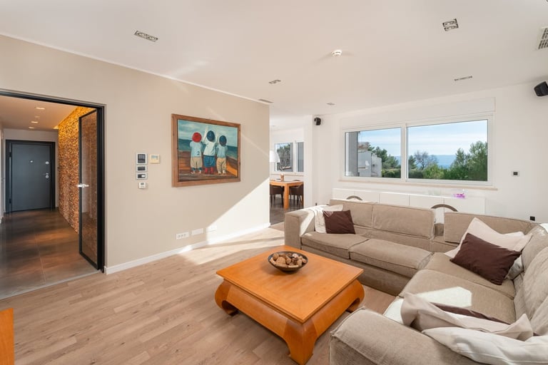 Modern living room with beige L-shaped sofa, wooden coffee table, artwork on walls, and large windows with natural light