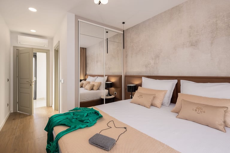 Modern bedroom with queen bed, mirrored wardrobe, beige bedding, wooden flooring, and green fabric accent piece