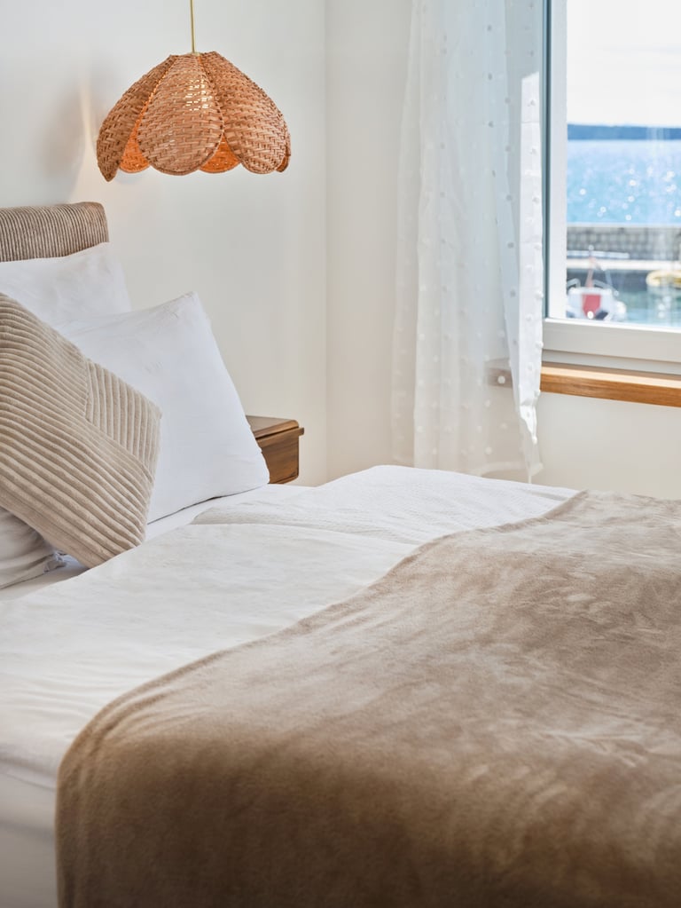 Minimalist bedroom with bed, white pillows, beige throw blanket, woven pendant lamp, and large window overlooking cityscape