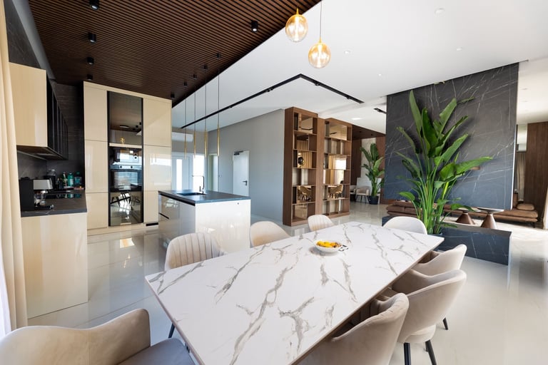 Modern open-plan kitchen and dining area with marble dining table, cream chairs, light wood cabinetry, and potted plant