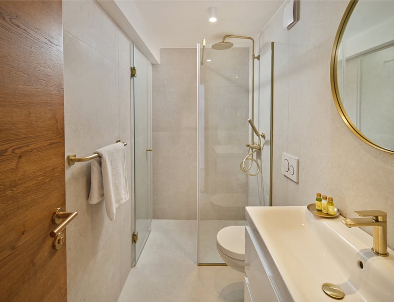 Modern bathroom with glass-enclosed shower, white sink, toilet, and gold-framed mirror on cream-colored walls