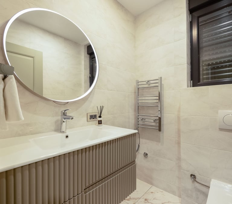 Modern bathroom with circular illuminated mirror, white sink, ribbed vanity cabinet, heated towel rack, and neutral tile walls