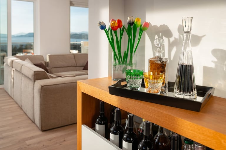 Modern apartment interior with wooden table displaying colorful tulips, decorative bottles, and glasses overlooking city views and beige sectional sofa