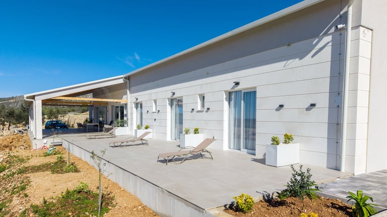 Modern white single-story house with covered patio, lounge chairs, and concrete deck under clear blue sky