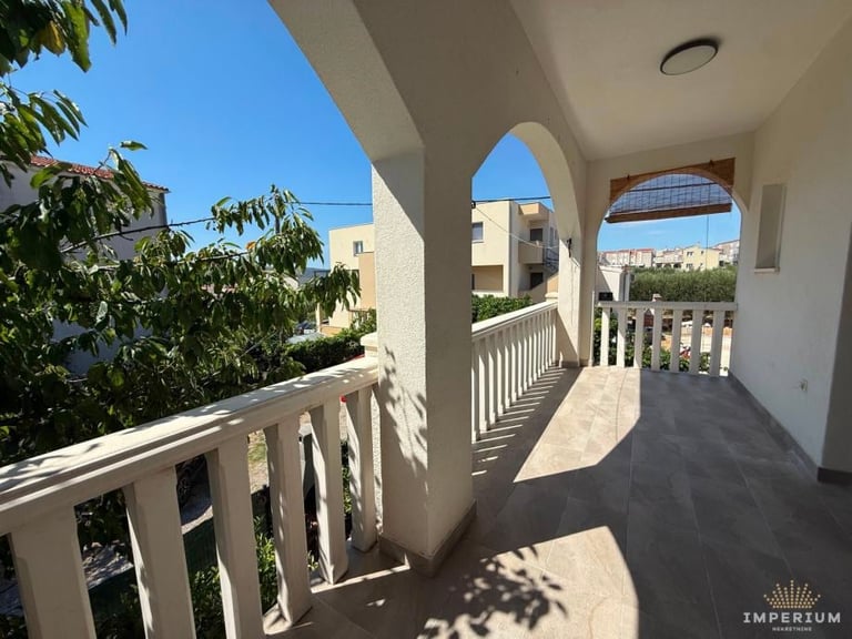 Sunny Mediterranean terrace with white railings, arched columns, and sea views. Mediterranean buildings visible in distance.