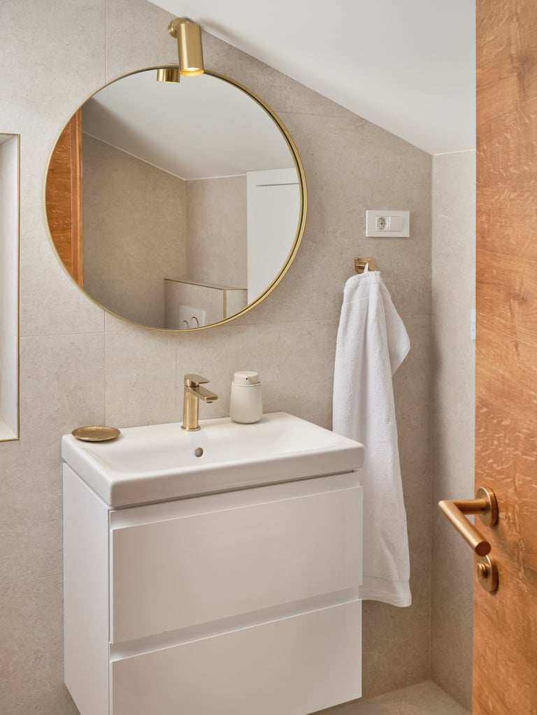Modern bathroom with white vanity, circular brass-framed mirror, gold fixtures, and beige tile walls with wooden door frame