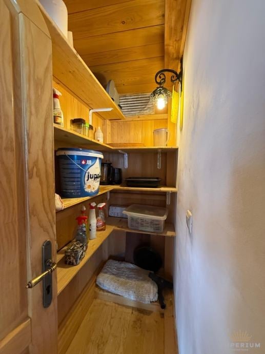 Narrow wooden storage closet with shelves containing household items, containers, and supplies, illuminated by warm lighting.