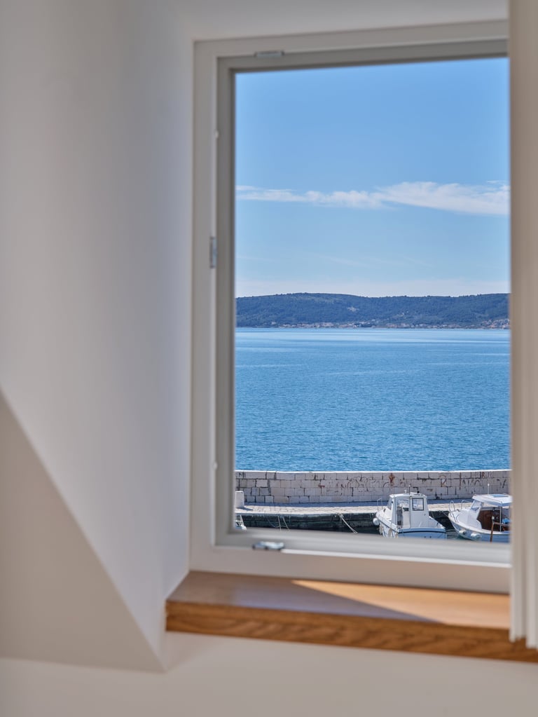 Window view of calm blue ocean and distant hills with boats along a dock, white interior walls visible